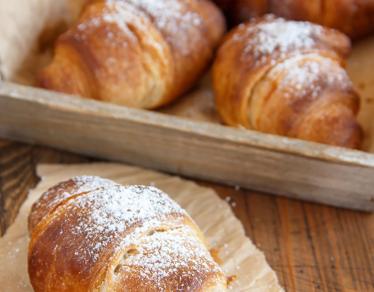 buttery croissant dusted with powdered sugar on parchment, with a wooden tray of croissants in the background