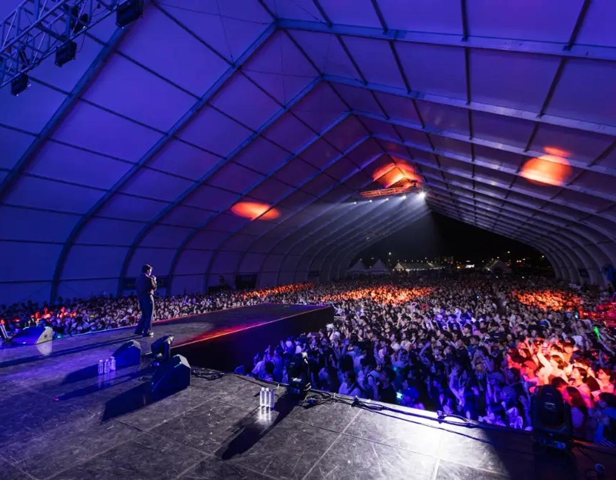 Covered outdoor concert stage with a solo performer facing a packed, colorfully lit crowd under a large arched tent