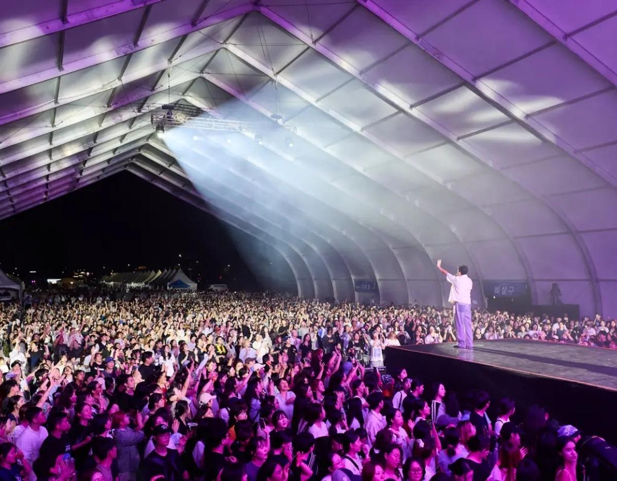 Large nighttime K-pop festival crowd under a lit canopy, performer on elevated stage facing thousands of cheering fans.