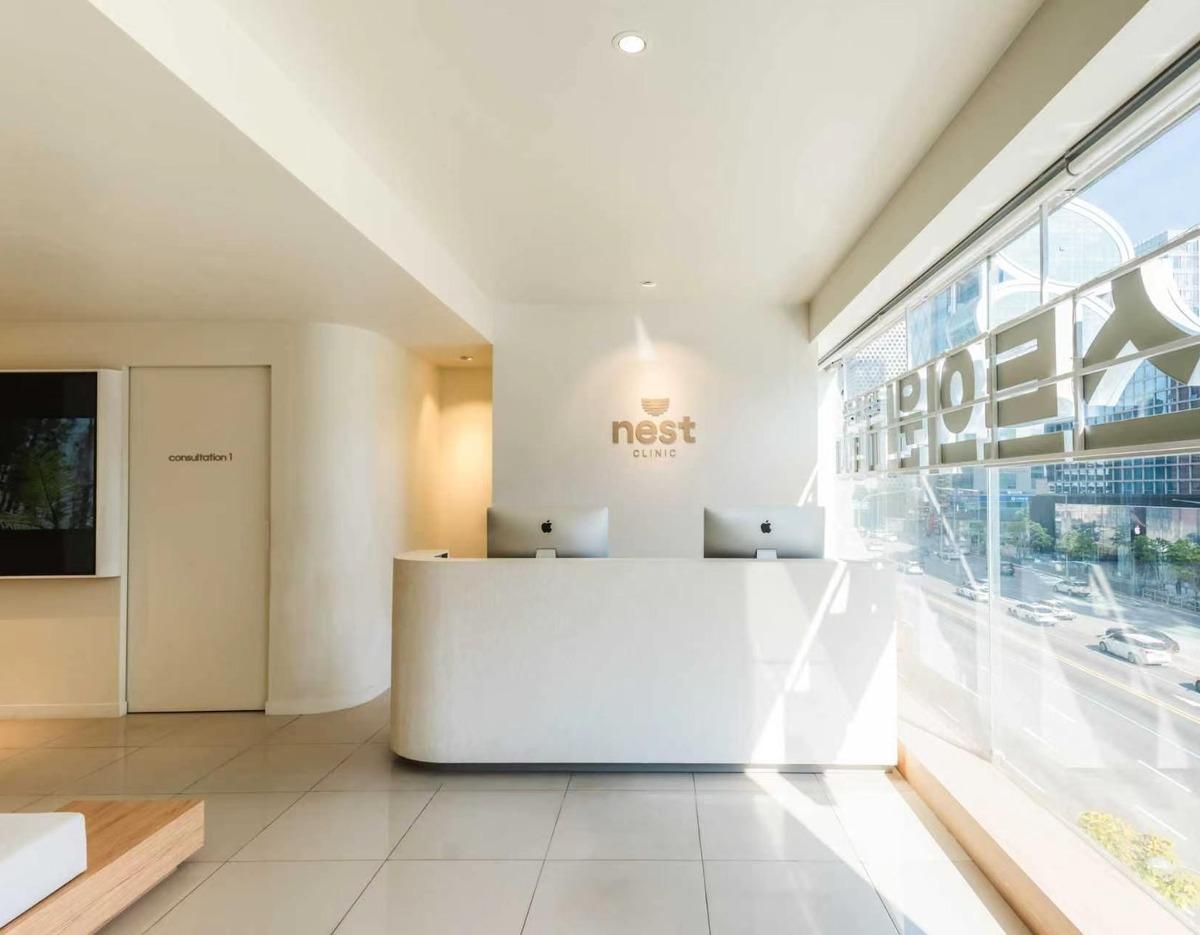Bright minimalist clinic reception with curved white desk, two iMacs, "nest clinic" logo on wall and large street-facing window.