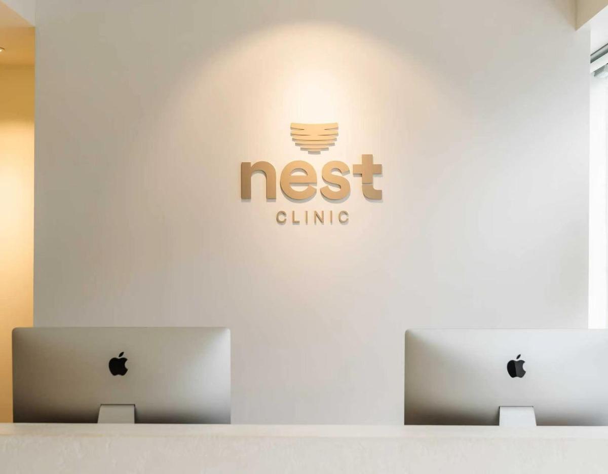 Minimal reception desk with two iMacs and a gold "nest CLINIC" logo on a white wall.