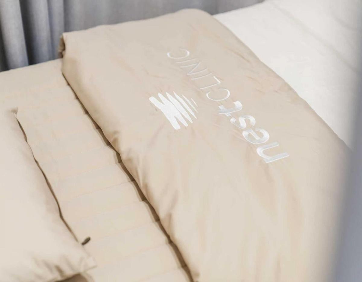 Beige treatment bed with embroidered "Nest Clinic" logo on a padded sheet in a calm consultation room.