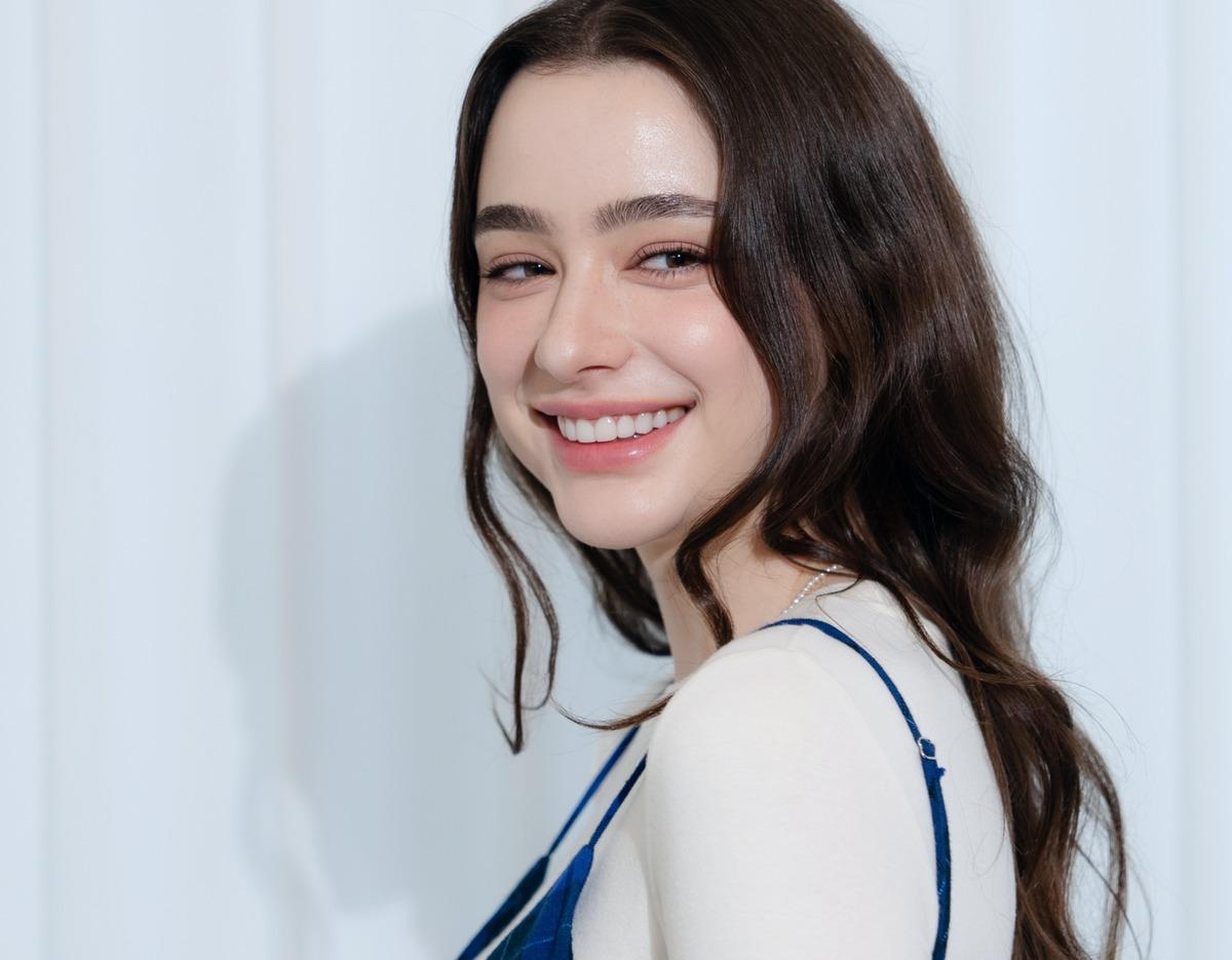 young woman with long wavy brown hair smiling brightly showing straight white teeth, wearing white top and blue straps