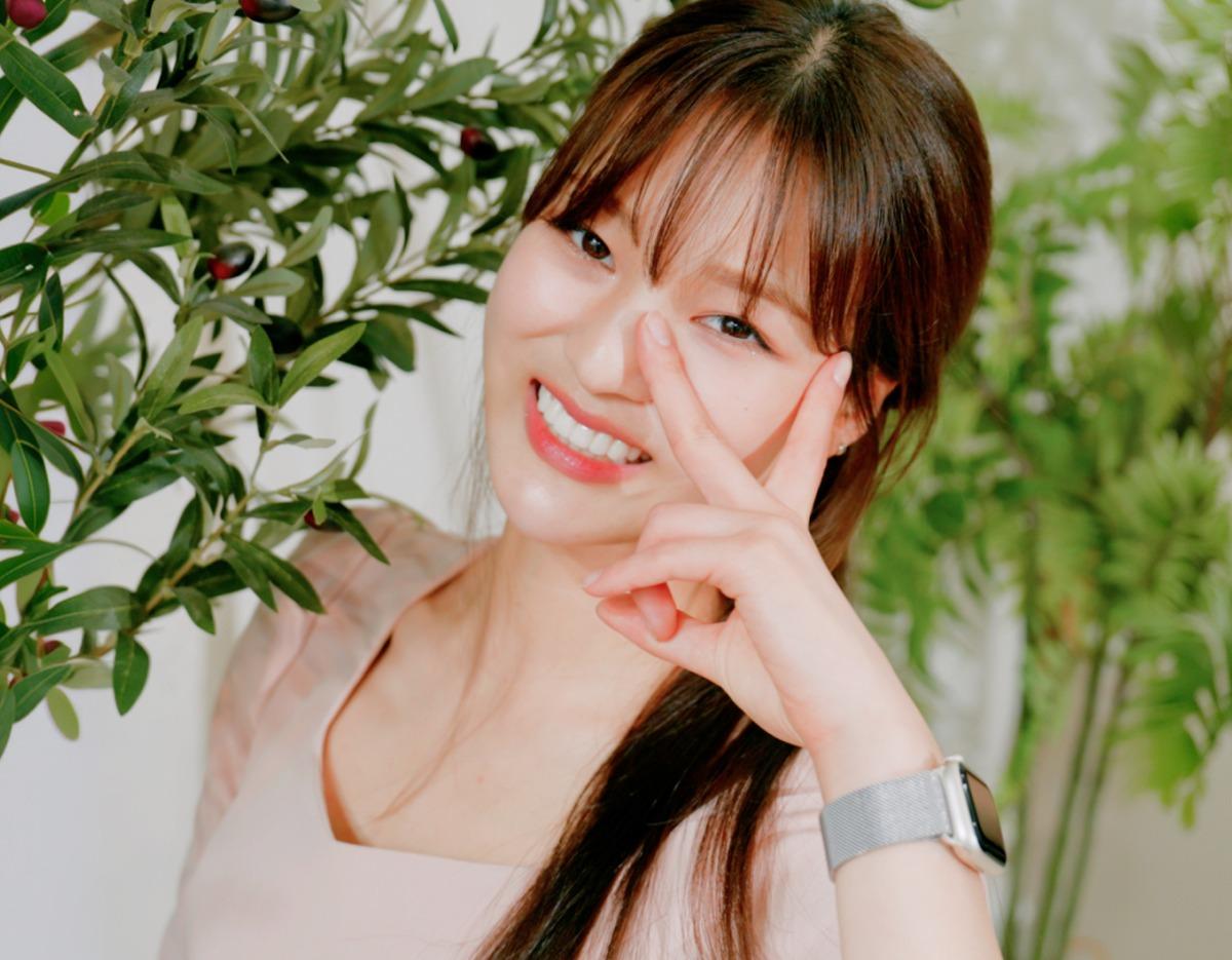 young woman smiling showing teeth with V sign near eye, wearing pink top and smartwatch, leafy plants background