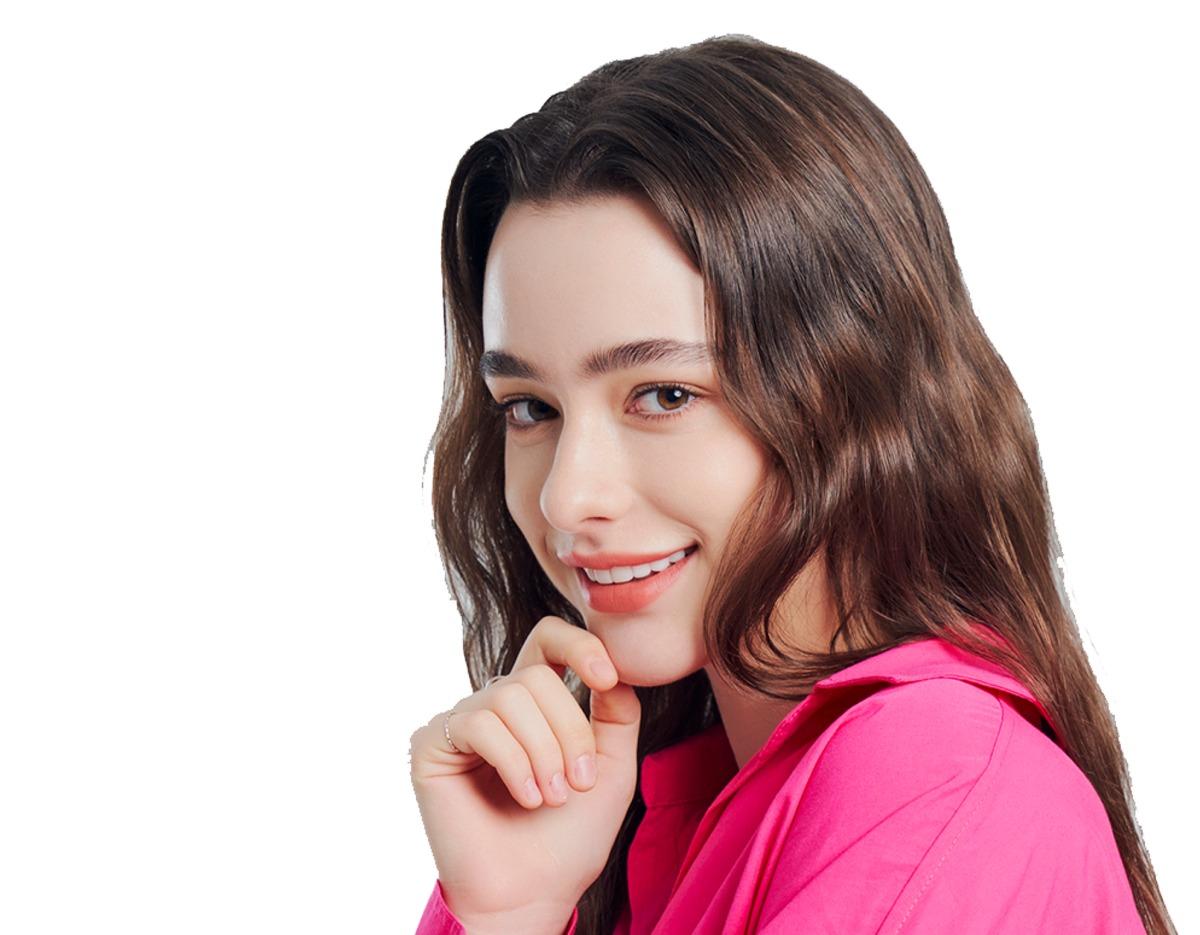 Young woman with long wavy brown hair smiling, wearing a bright pink collared shirt and resting chin on her hand.