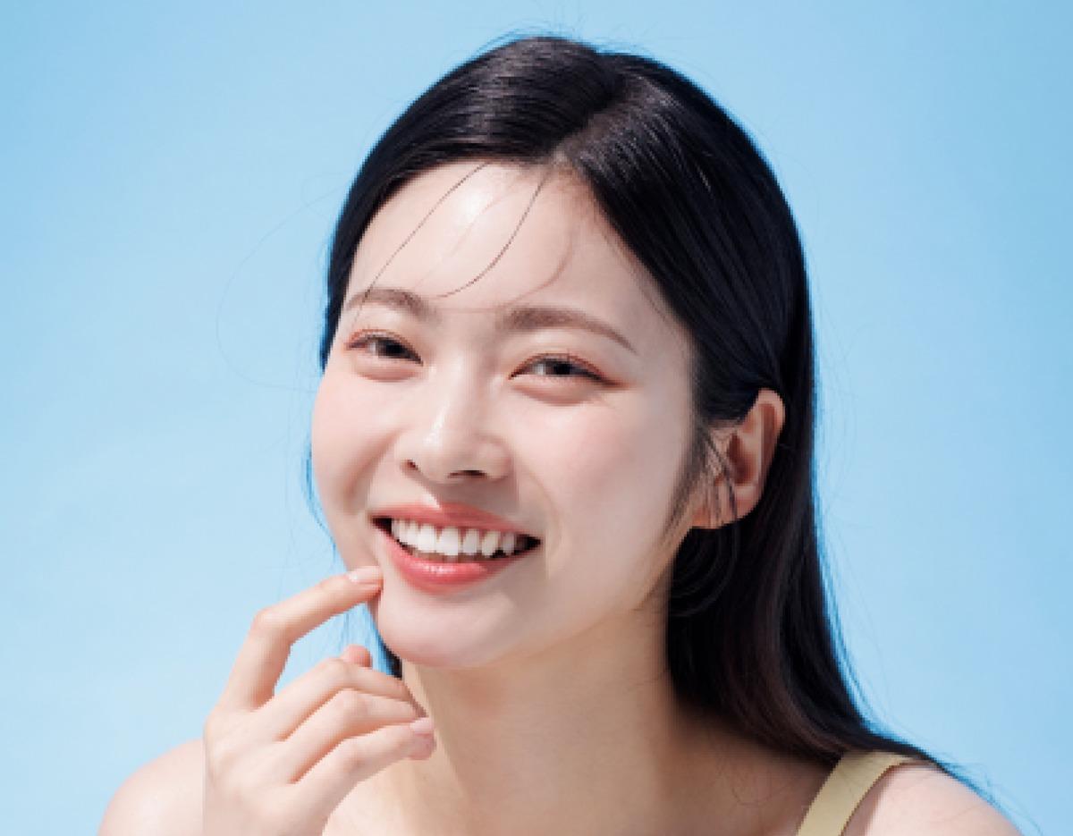 young woman with long dark hair smiling against a pale blue background, hand touching chin showing white teeth