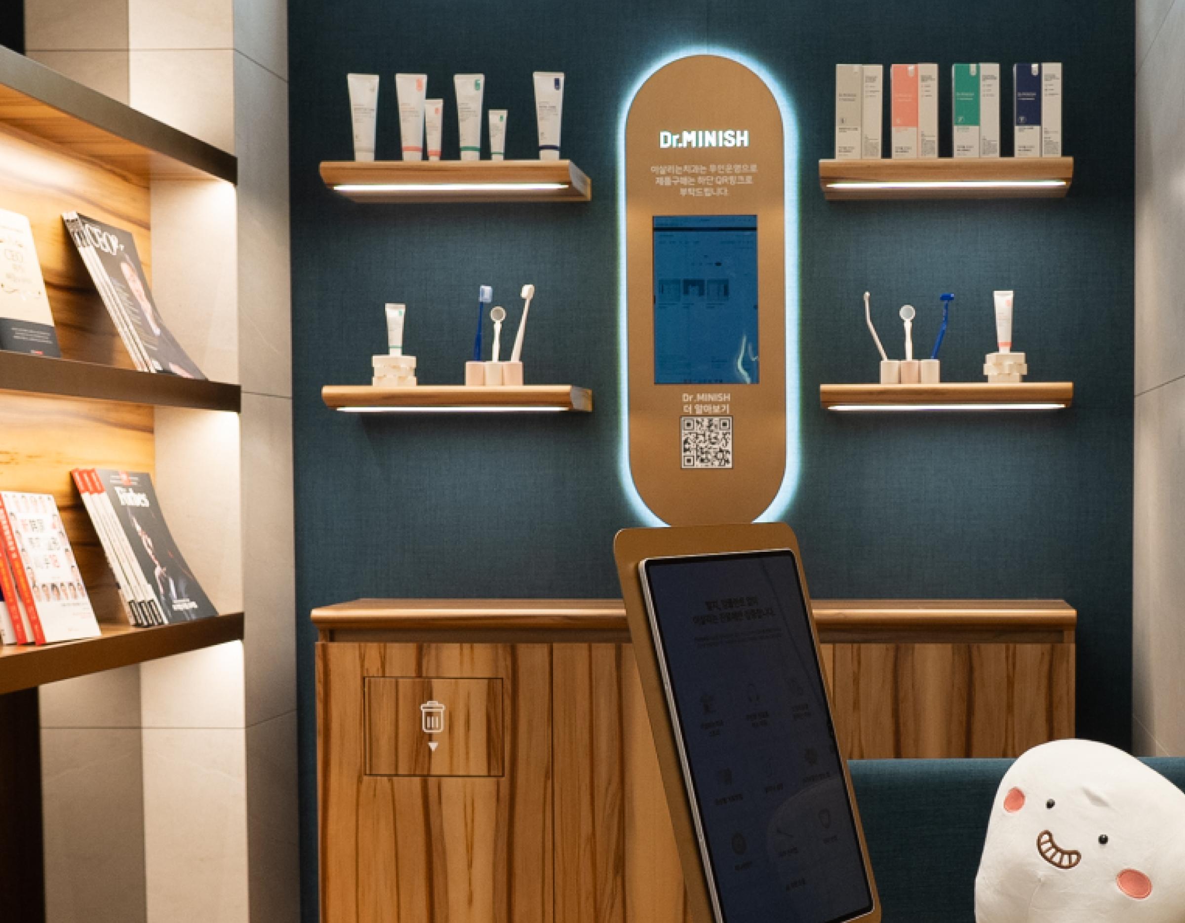 Reception display wall with wooden shelves holding dental products, a lit Dr.MINISH sign and a tablet kiosk in clinic waiting area