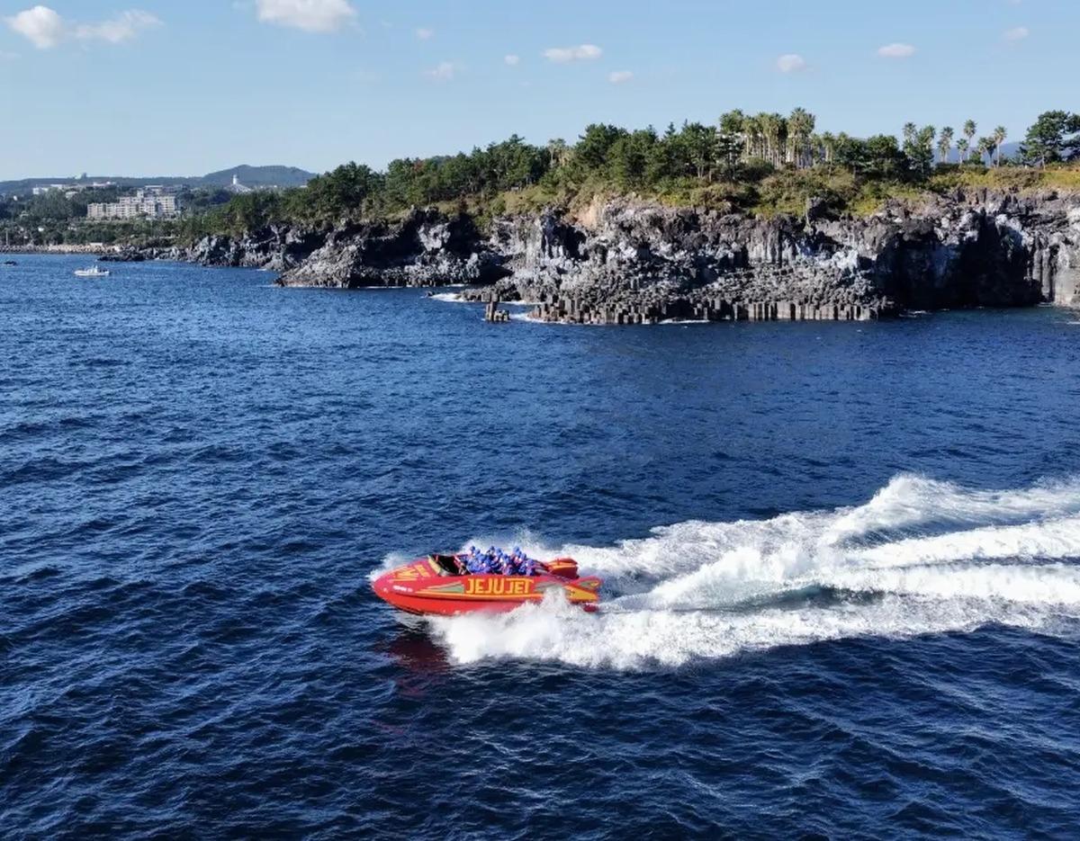 red JEJU JET speedboat with passengers racing across blue sea past rocky Jusangjeolli coastal cliffs under a sunny sky