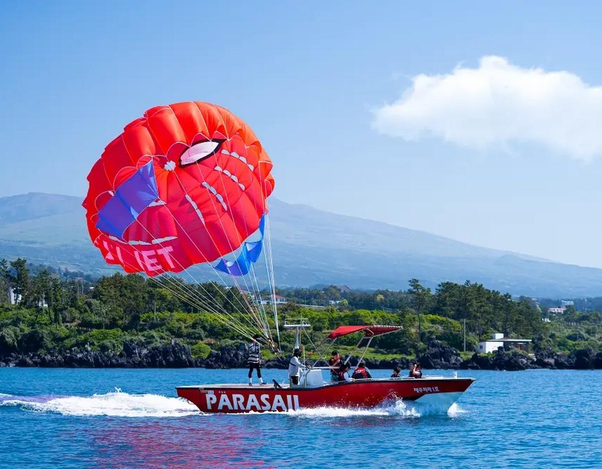 Red motorboat labeled "PARASAIL" towing a bright red parachute with passengers over blue sea near rocky Jeju coastline.