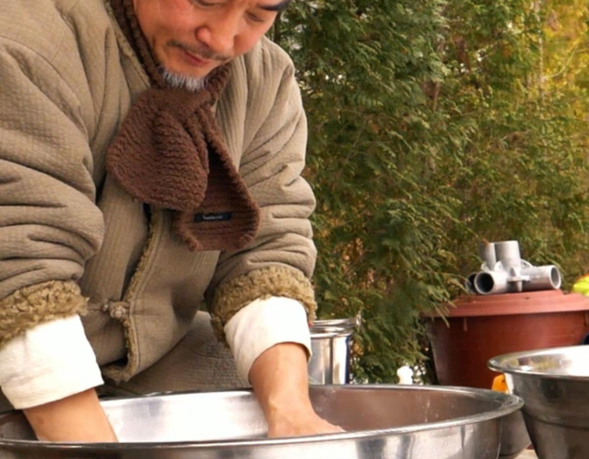 Man in warm jacket and scarf kneading dough or batter in a large metal basin outdoors near greenery and cooking pots