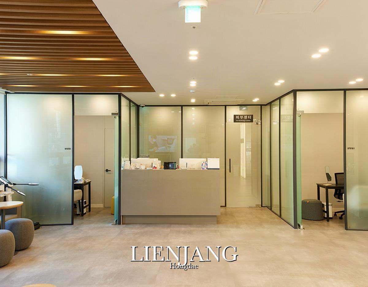 Modern clinic reception with central desk, frosted glass consultation rooms, wood ceiling slats and lounge seating at 리엔장의원 홍대점