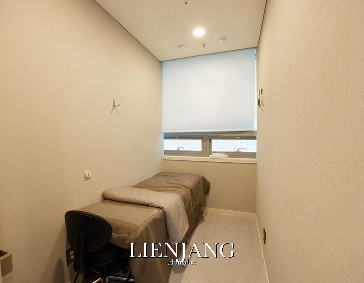 Small private treatment room with single covered bed, rolling chair, window with blind and neutral beige walls at 리엔장의원 홍대점.