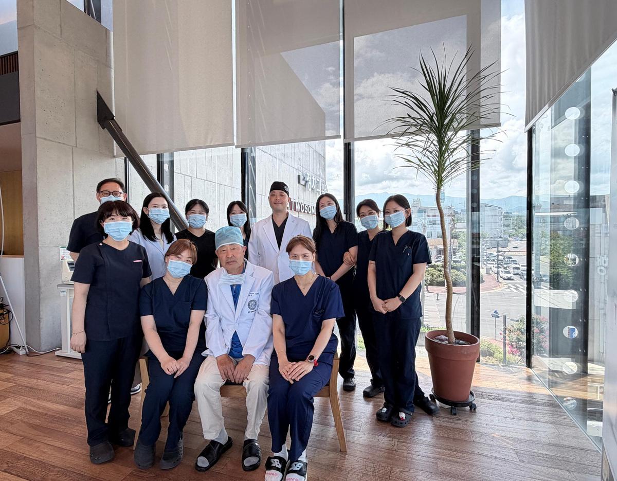 Dental clinic team of doctors and staff in scrubs and masks posing by large glass windows with potted plant.