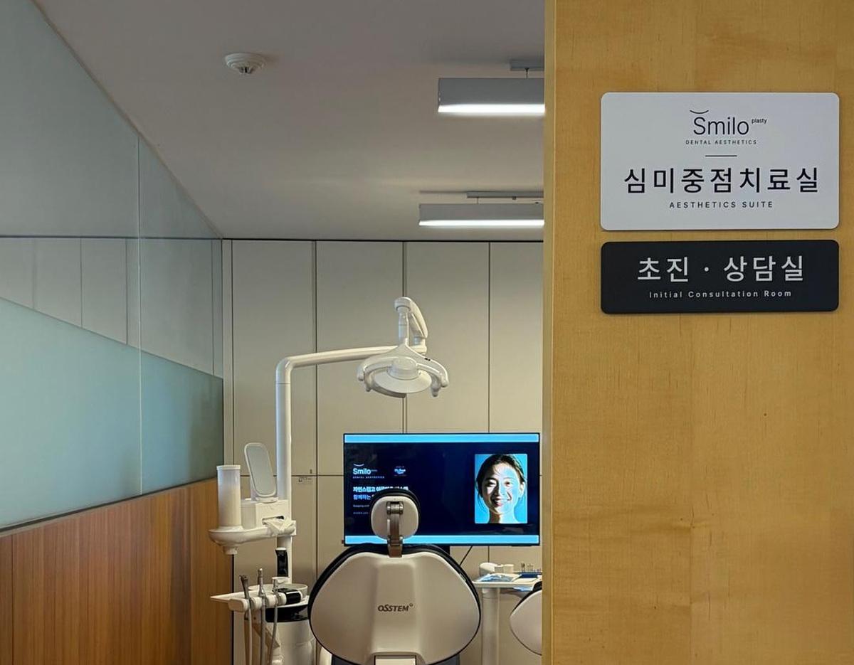 Dental treatment room with Osstem dental chair, overhead lamp, monitor showing a smiling face, and door signs reading "Aesthetics Suite" and "Initial Consultation Room