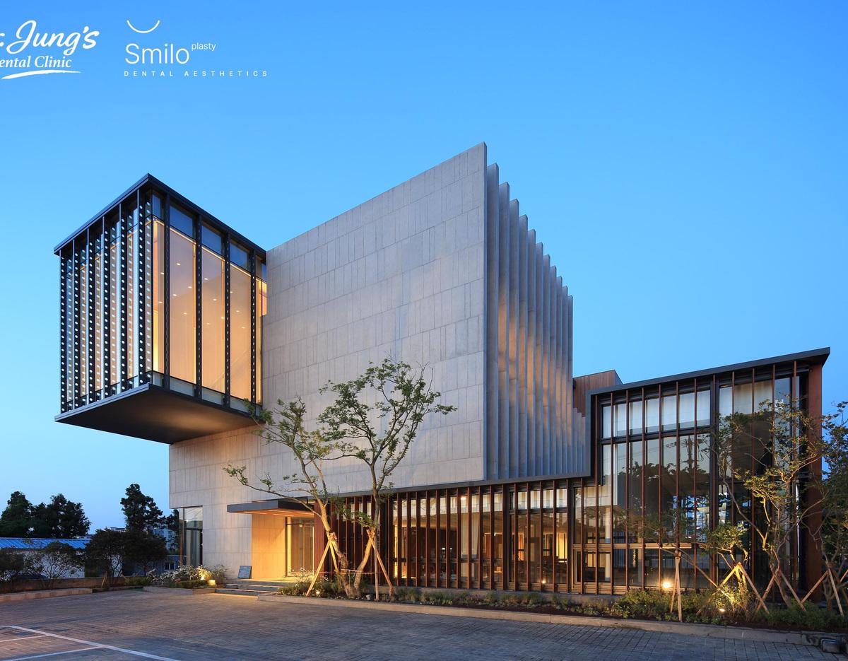Modern multi-story dental clinic building at dusk with cantilevered glass box, tall concrete facade and landscaped entrance.