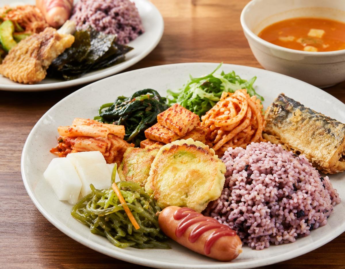Korean buffet plate with purple multigrain rice, sausage with ketchup, fried fish, pancakes, kimchi and assorted banchan sides