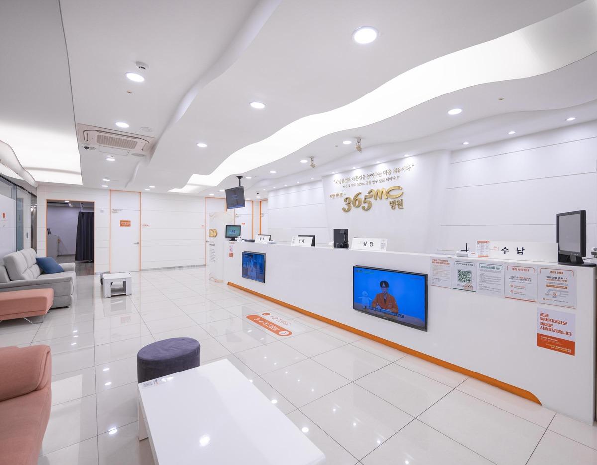 365mc병원 부산점 bright white reception area with long desk, seating, wall logo and TV screens