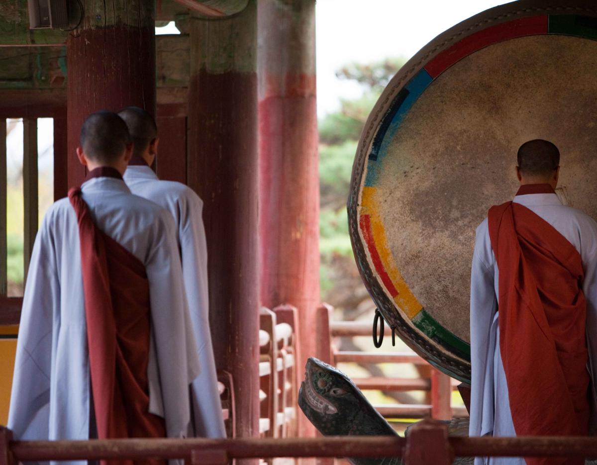 Three Buddhist monks in white robes and red shoulder cloaks standing by a large painted temple drum on a wooden pavilion