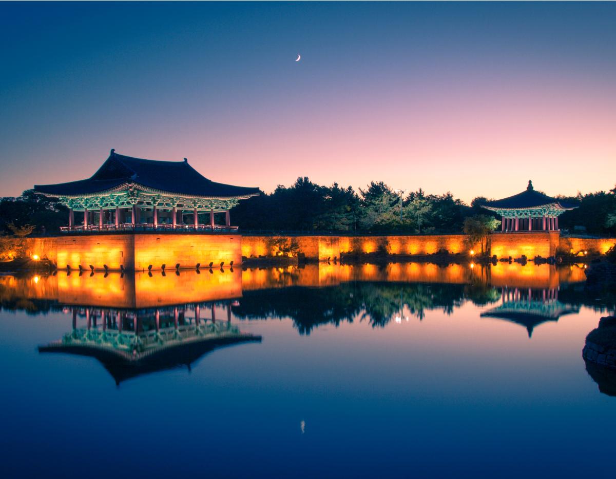 Gyeongju illuminated traditional pavilion and fortress wall reflected in calm pond at twilight with crescent moon