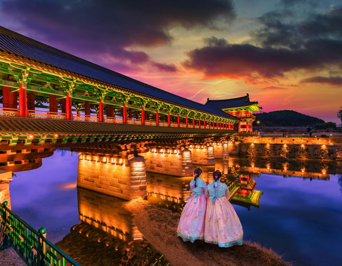 Illuminated traditional covered bridge over calm water at dusk with two women in pastel hanbok looking across reflections
