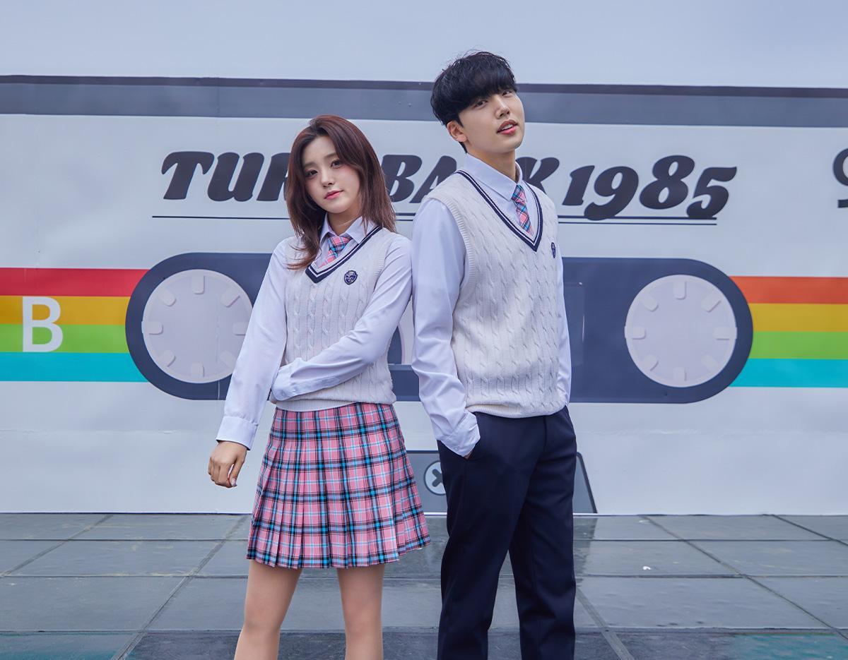 two young models in matching school uniforms—white cable-knit vests, white shirts and pink plaid skirt and tie—posed in front of a retro cassette mural