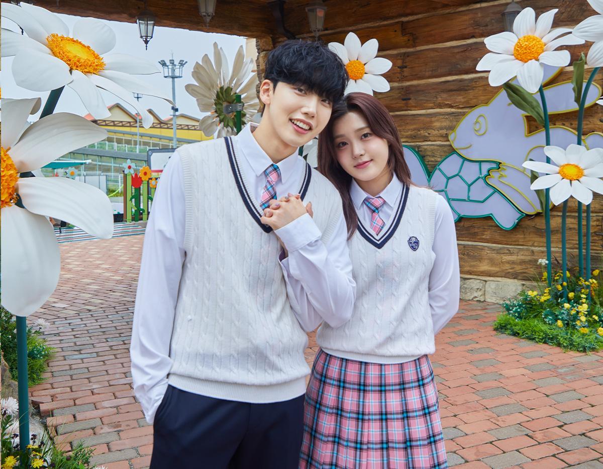 two young people in matching white cable-knit vest school uniforms and pink plaid ties posing amid oversized daisy decorations