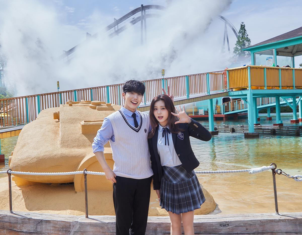 young man and woman in matching Korean school uniforms posing by water and amusement park ride at Gyeongju World