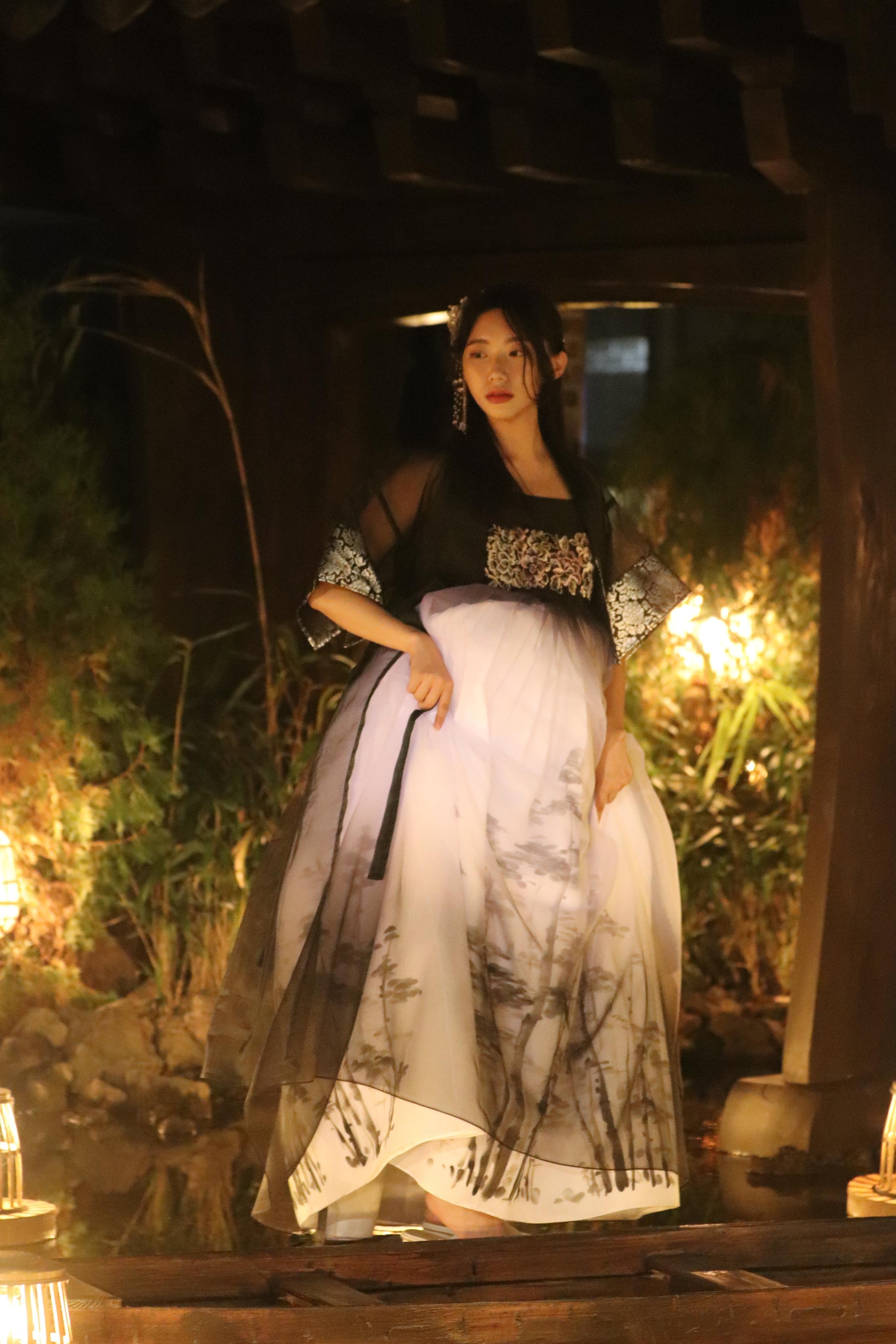 Woman wearing a flowing black-and-white hanbok standing on a wooden bridge at night, lit by warm lanterns