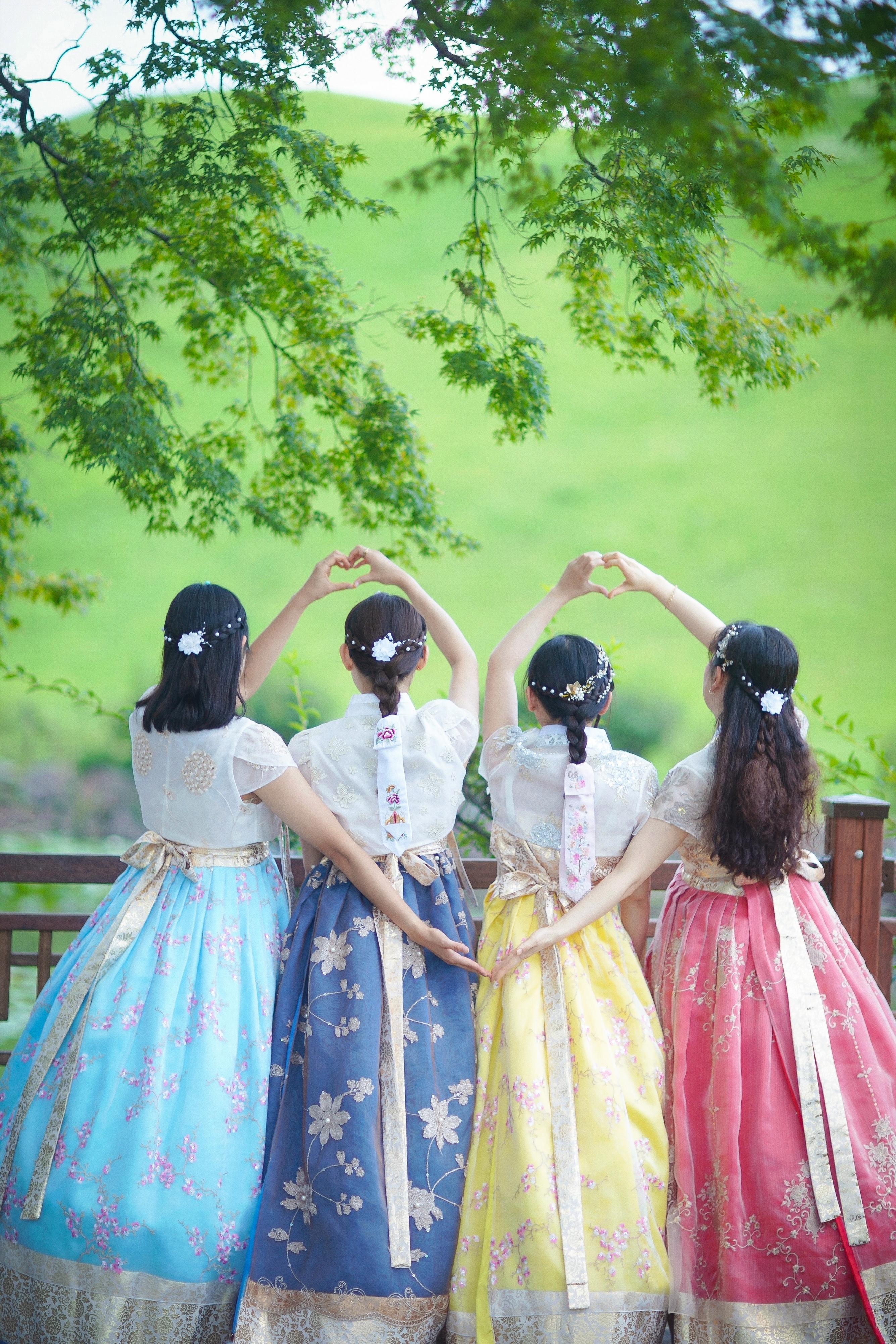 four women in colorful hanbok (blue, navy, yellow, pink) stand with backs to camera under green trees, making heart shapes with hands