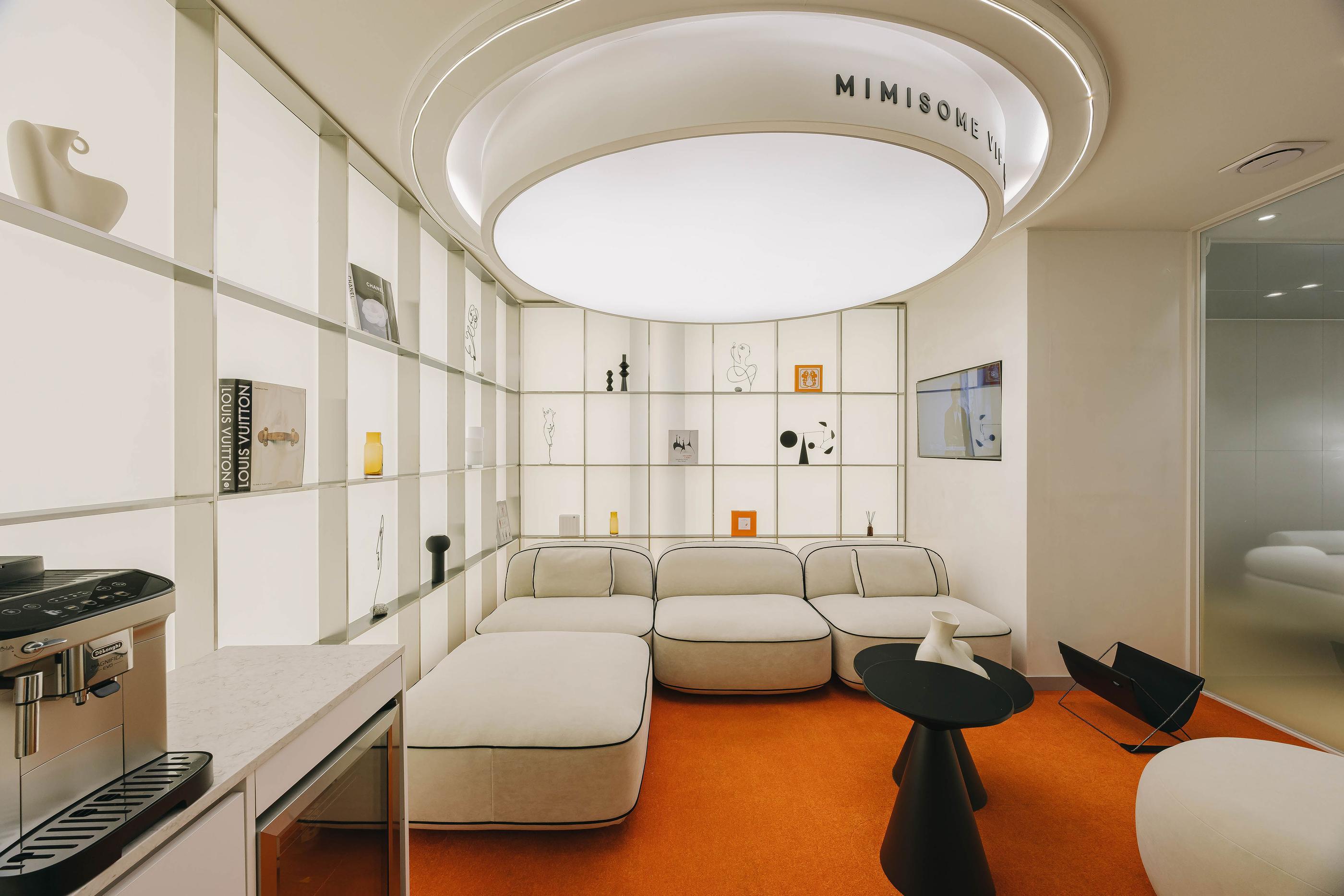 MIMISOME clinic lounge with curved white modular sofa, round overhead light fixture, orange carpet and coffee machine.