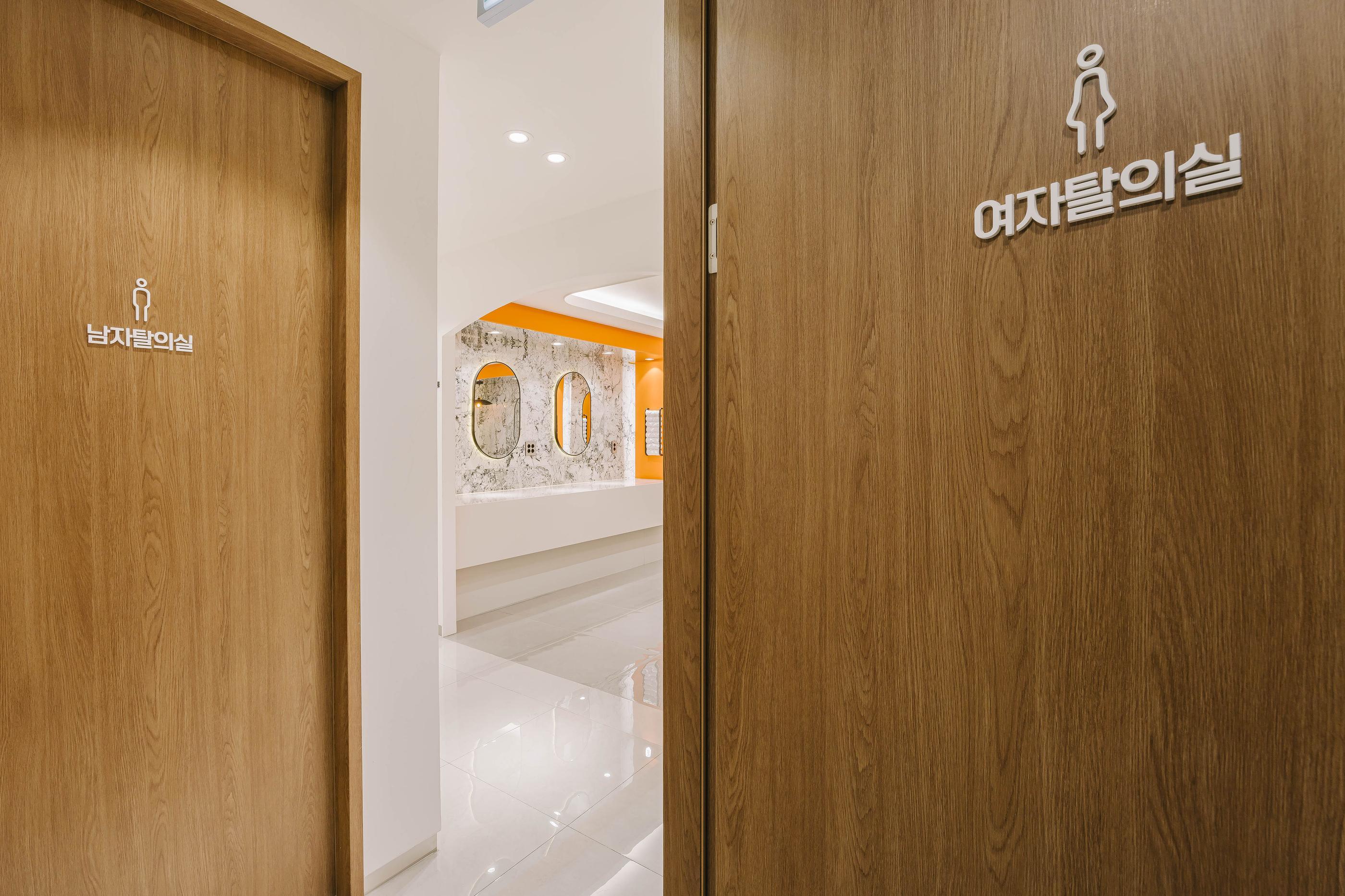 Clinic hallway showing wooden restroom doors labeled in Korean for men and women, with view into bright vanity area with mirrors.
