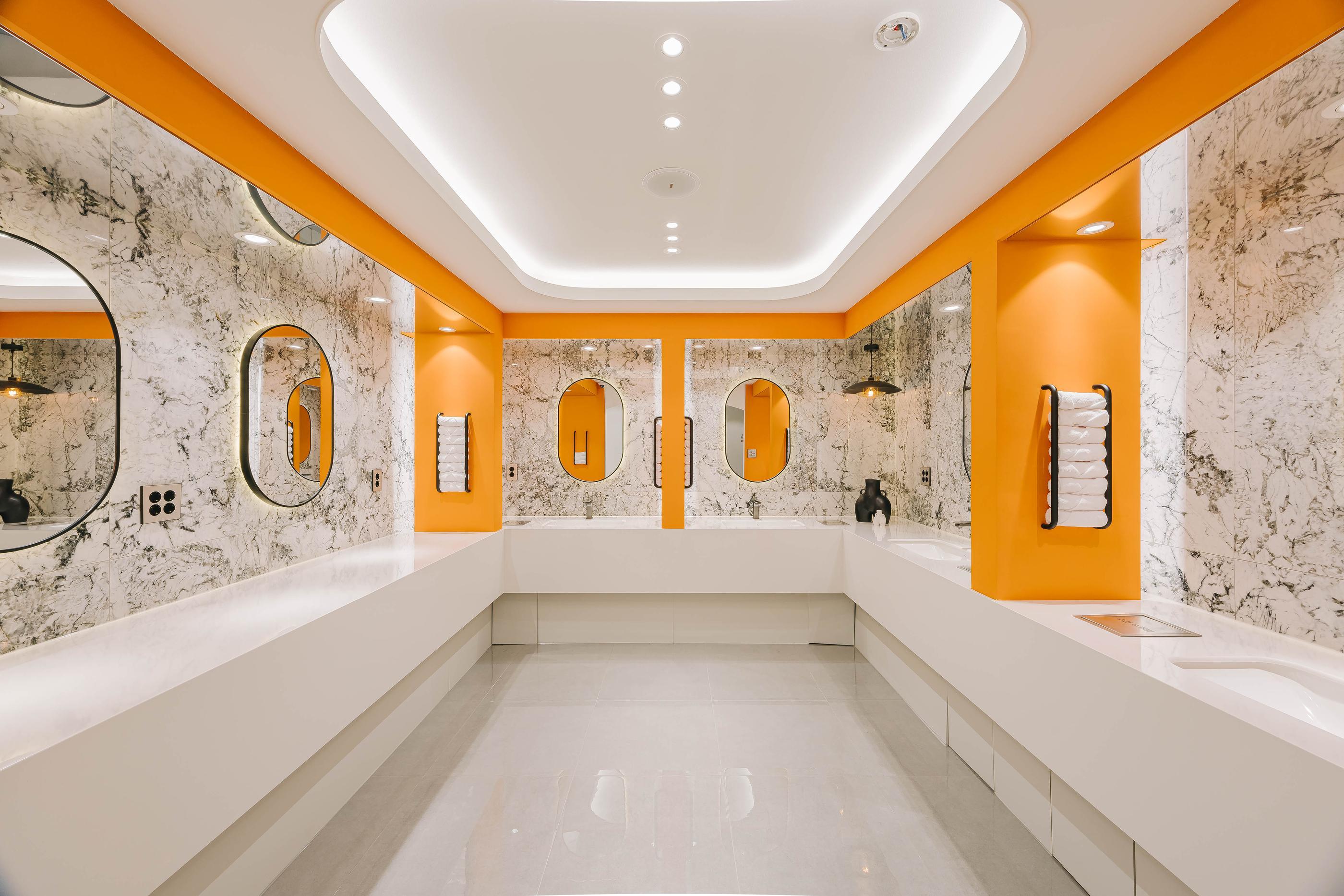 Bright clinic vanity area with orange accents, marble walls, oval mirrors and long white countertops at 미미썸의원 부산덕천점