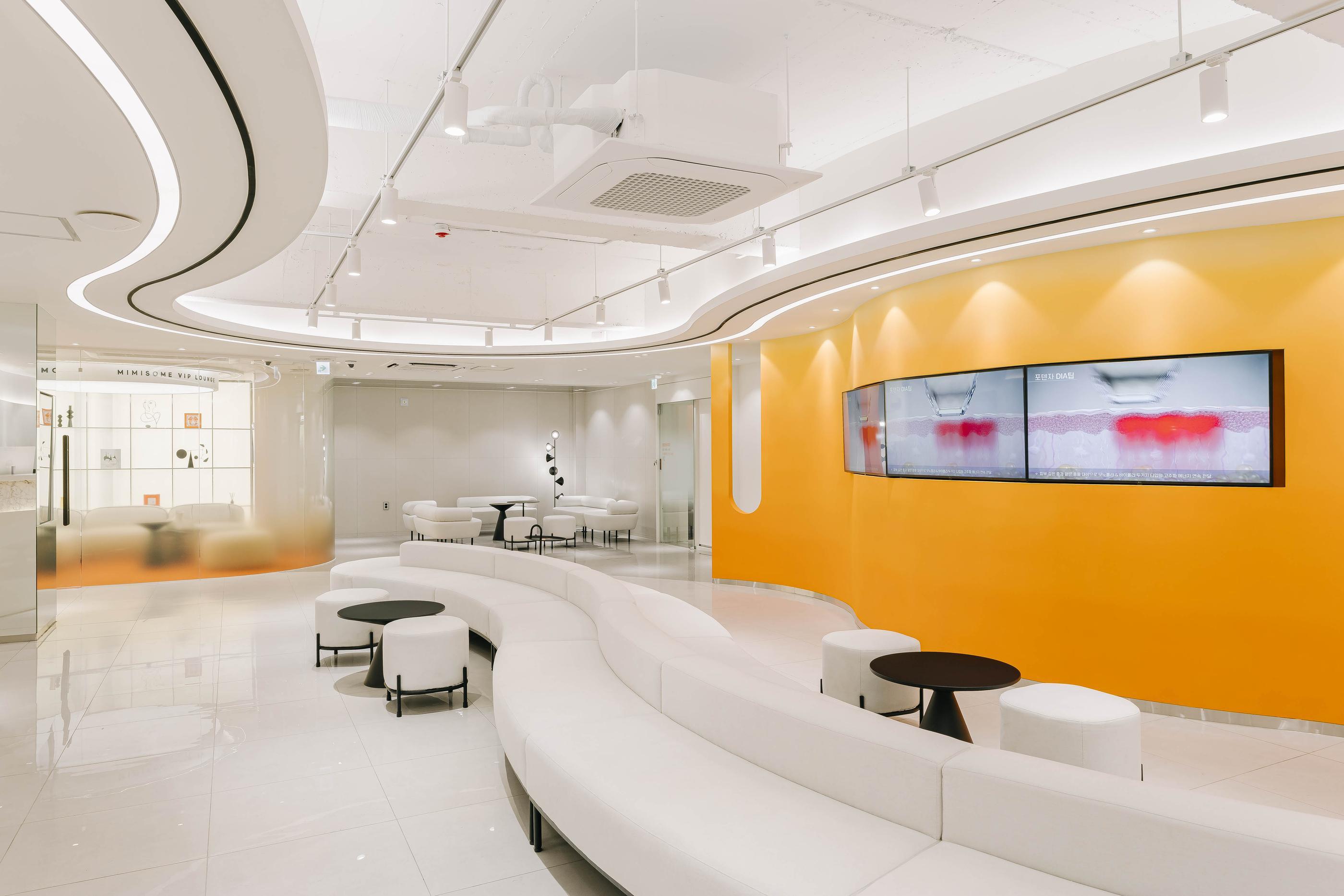 미미썸의원 부산덕천점 bright curved lounge with long white modular sofa, round stools and yellow feature wall with screens