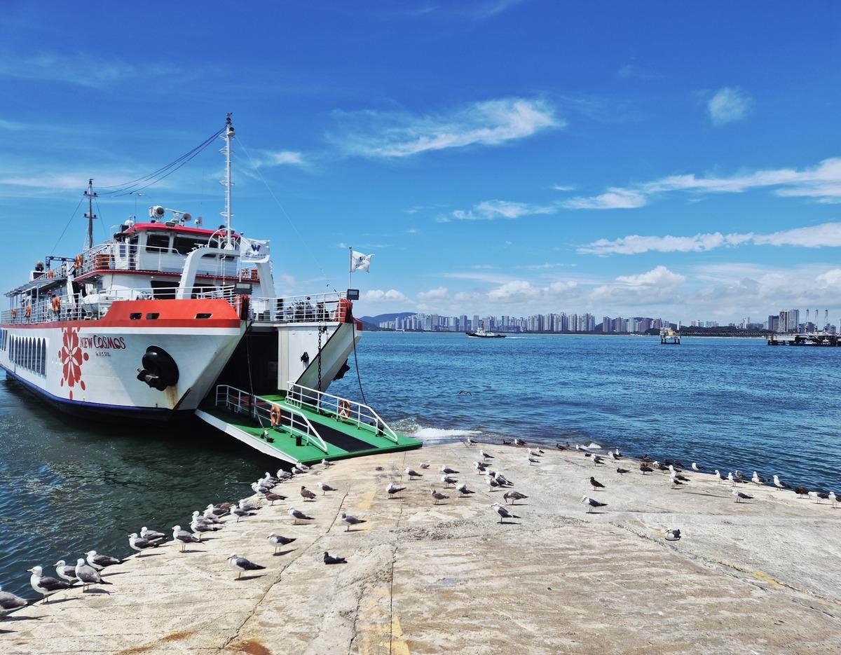 New Cosmos ferry docked at Wolmido with open ramp, seagulls on concrete pier and Incheon skyline across blue sea
