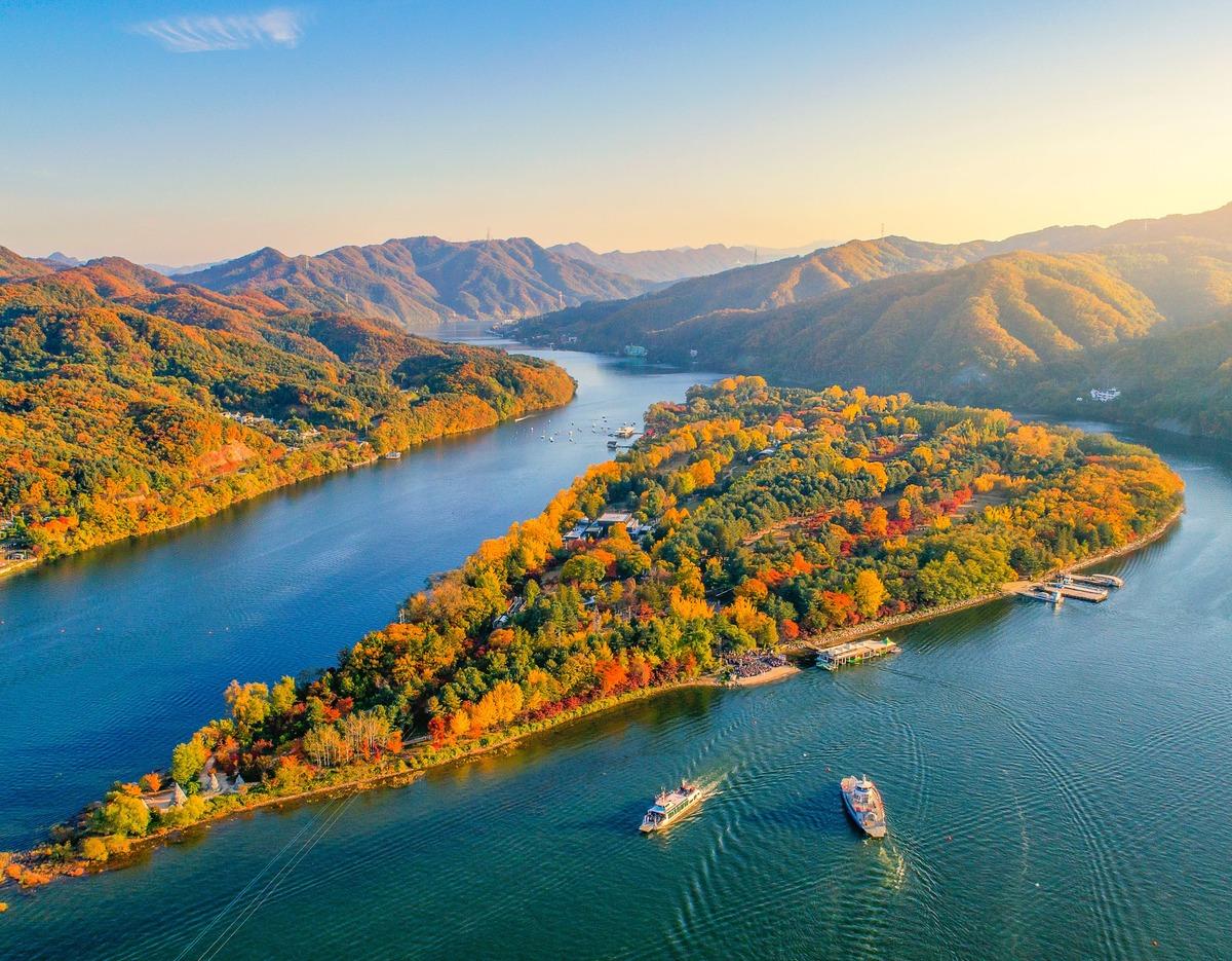 Nami Island aerial view in autumn with colorful trees, surrounding river, boats and forested mountains under golden light