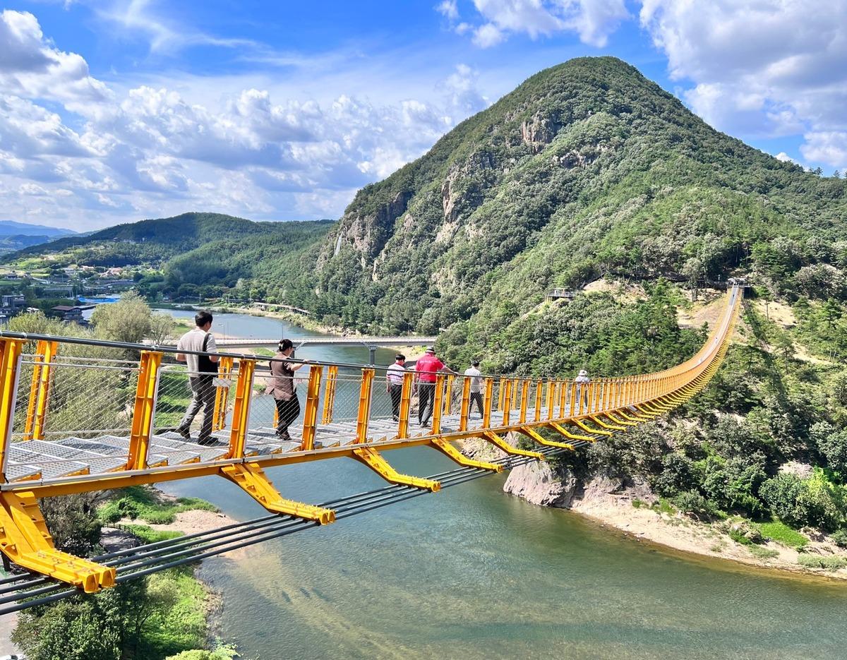 Yellow suspension footbridge crossing a river toward forested Wolyeongsan mountain with visitors walking on it.