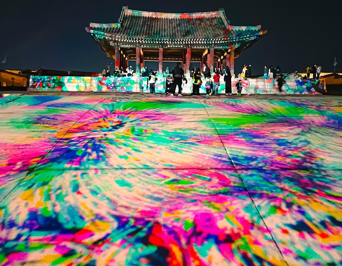 Suwon Hwaseong Festival nighttime scene: colorful projected light patterns on courtyard leading to illuminated traditional pavilion with visitors