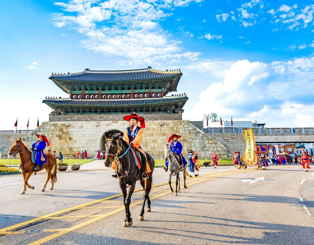 Horse-mounted performers in traditional Korean dress parading in front of Hwaseong Fortress gate under a blue sky