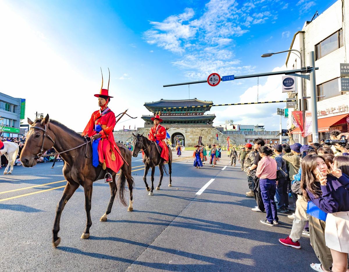Parade riders in red traditional hanbok on horseback passing Hwaseong Fortress gate with crowds watching along street