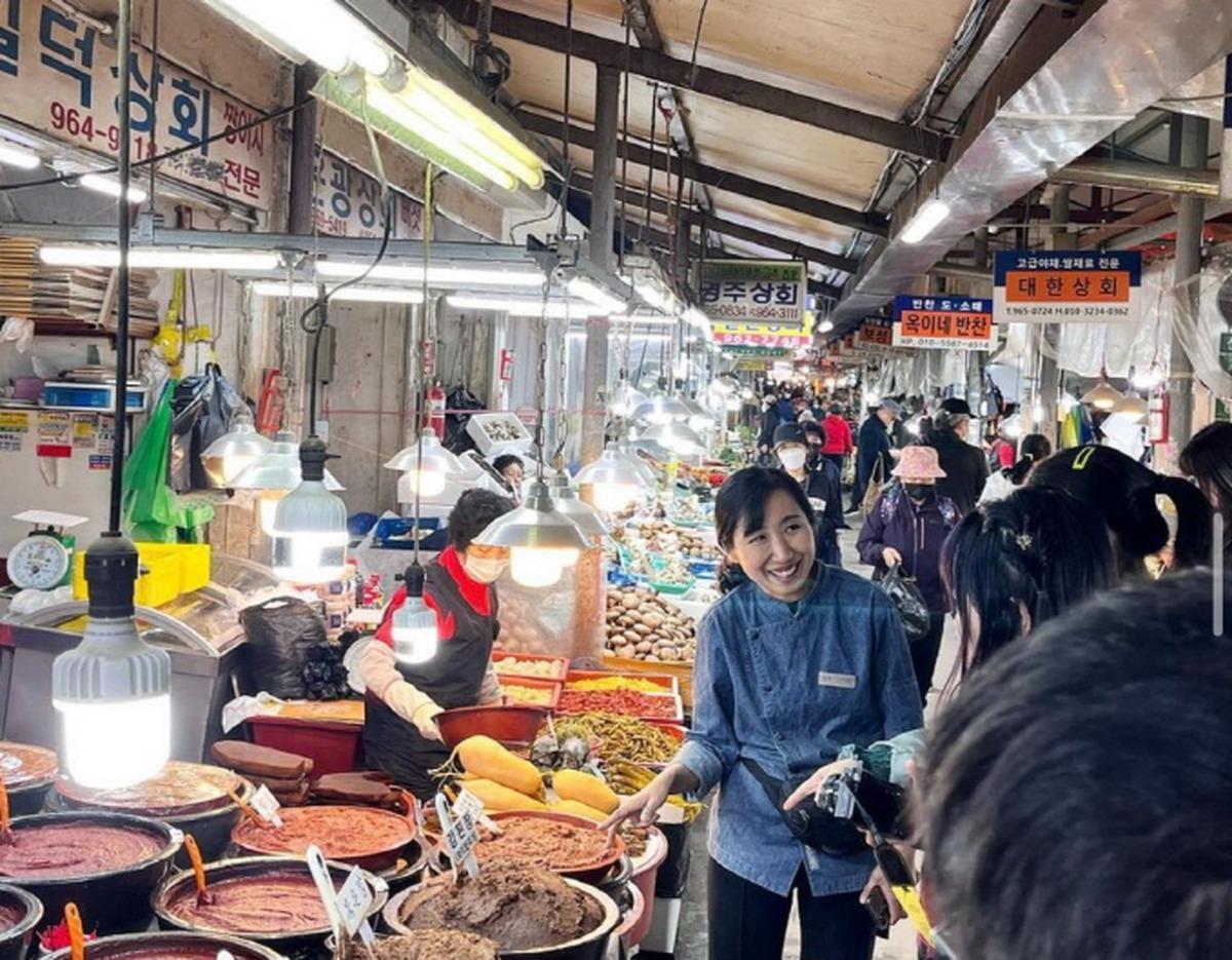 Busy indoor Korean market stall with a smiling instructor showing visitors tubs of red and brown pastes and fresh produce.