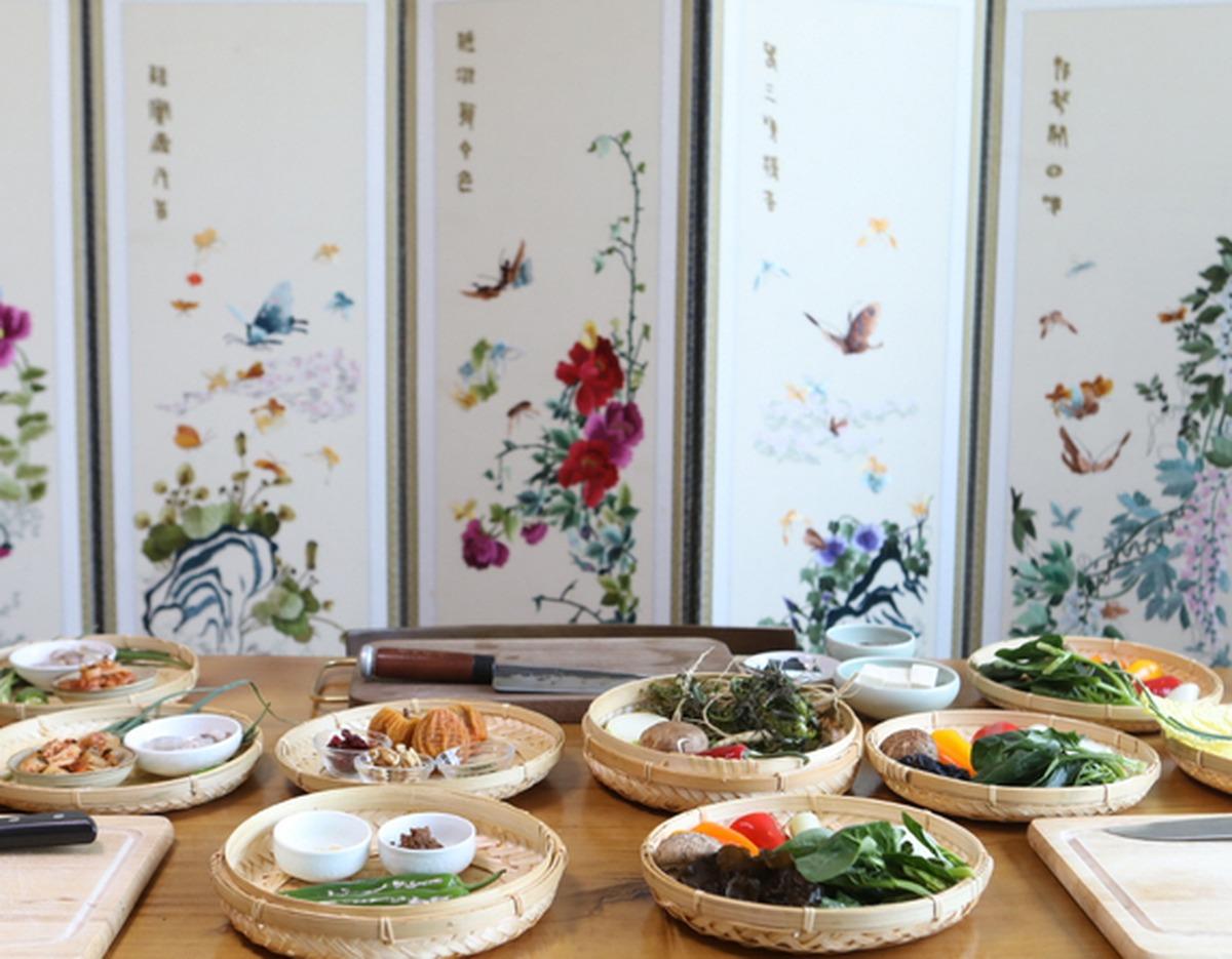 Traditional Korean cooking class setup with woven trays of vegetables, herbs, spices and small bowls on a wooden table, decorative folding screen backdrop.