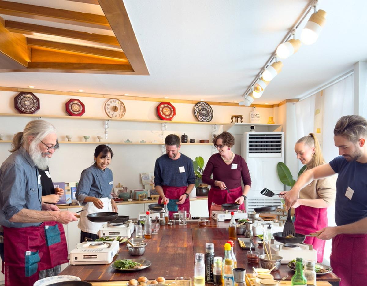 Participants wearing aprons cook at a long wooden table with portable stoves, pans, ingredients and smiling instructor.