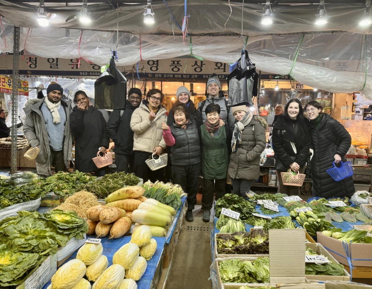 Group of diverse participants and vendors smiling at a traditional Korean market stall surrounded by fresh leafy greens and root vegetables.