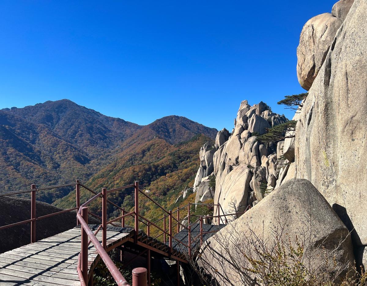 Wooden deck and metal railing along rocky ridge leading toward jagged Ulsanbawi granite peaks under clear blue sky in Seoraksan.