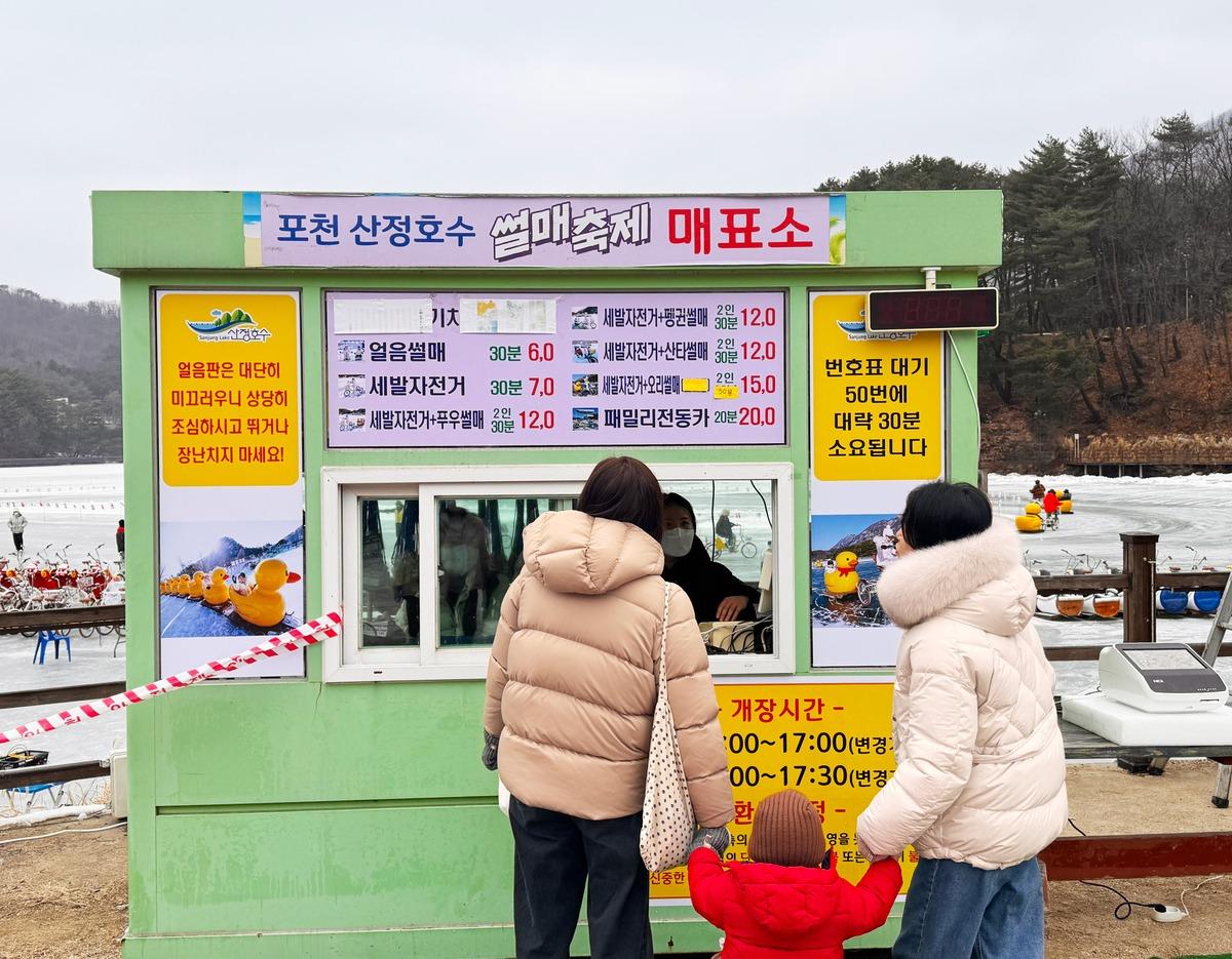 포천 겨울 일일 투어 ticket booth at frozen lake with two adults and child in winter coats buying tickets, rubber ducks on ice visible