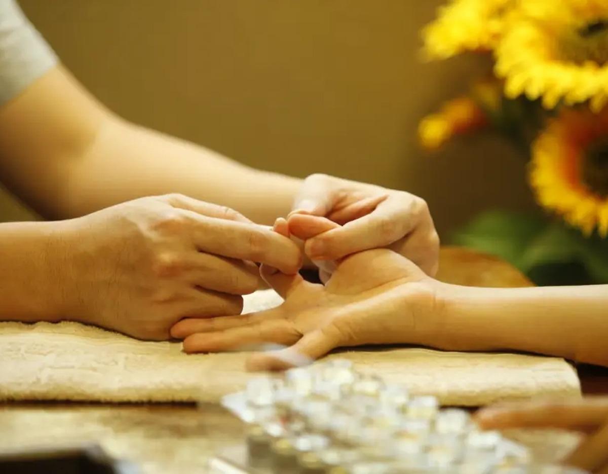 therapist's hands performing a gentle hand reflexology or pulse diagnosis on a client's palm with sunflowers blurred in background