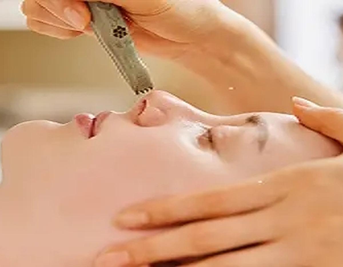 Client lying with eyes closed while therapist uses a metal 방짜괄사 tool to stimulate the nose and facial contours during treatment
