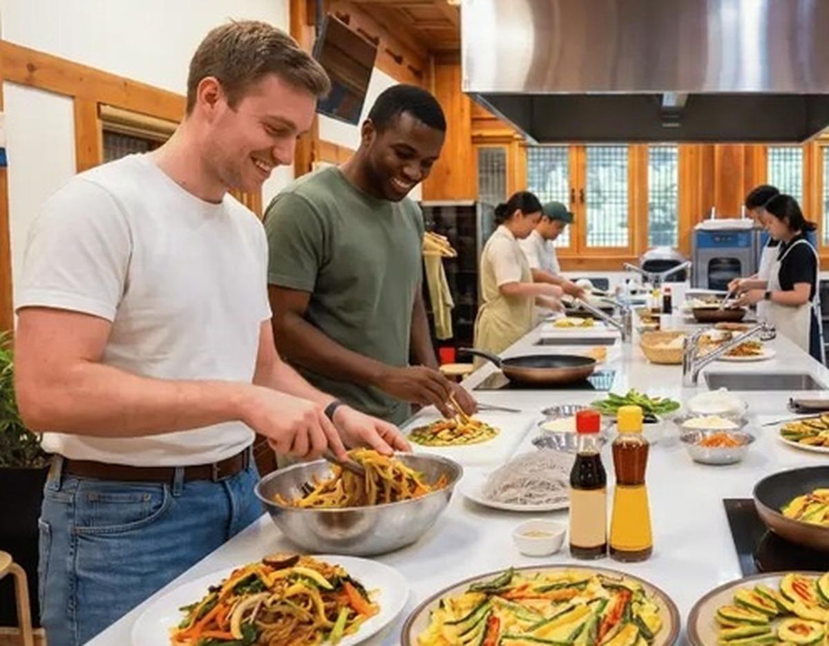 Foreign participants smiling as they plate homemade japchae and jeon at a long kitchen counter during a cooking workshop
