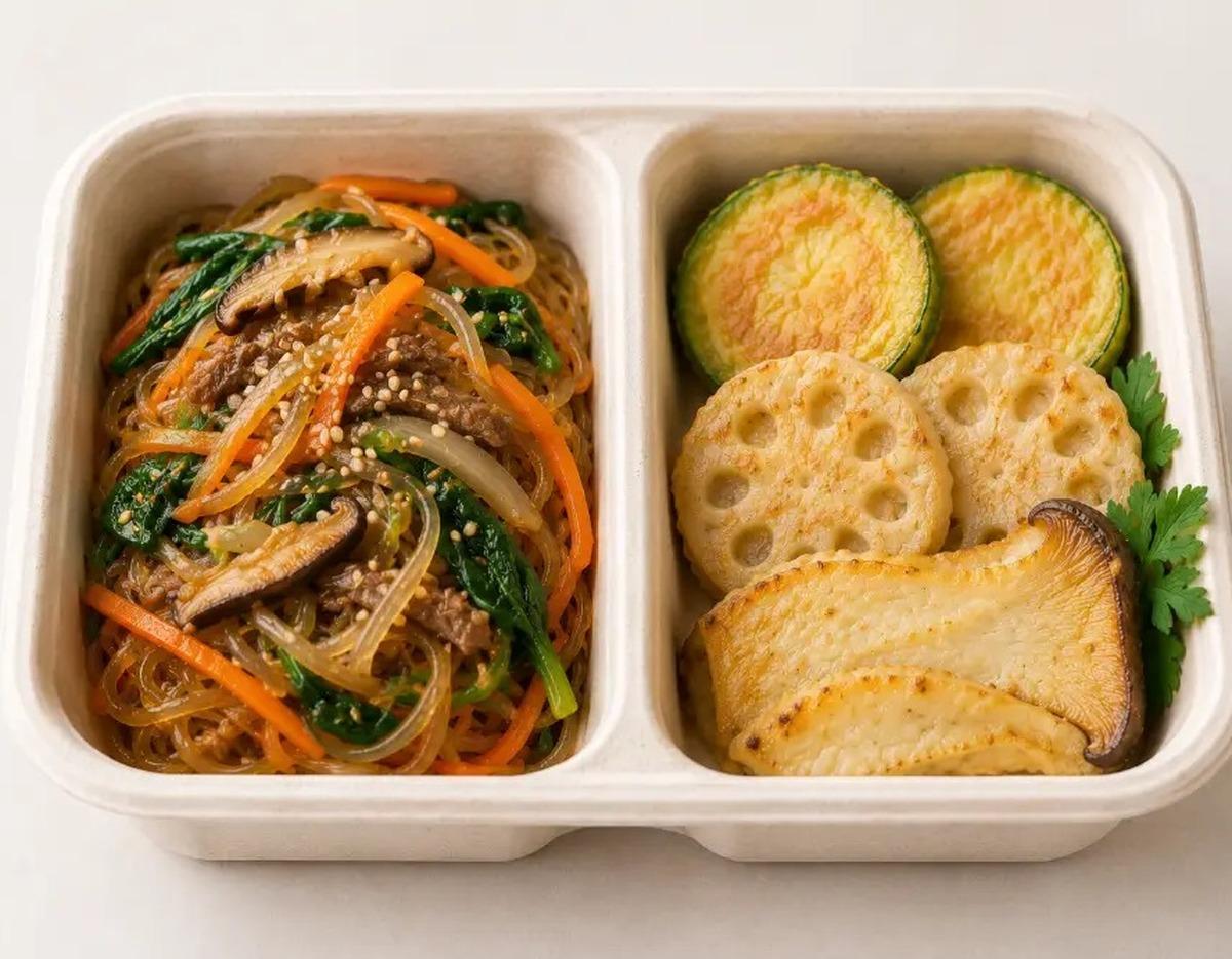 Bento-style tray with japchae noodles topped with mushrooms and vegetables, and pan-fried jeon slices (zucchini, lotus root, mushroom).