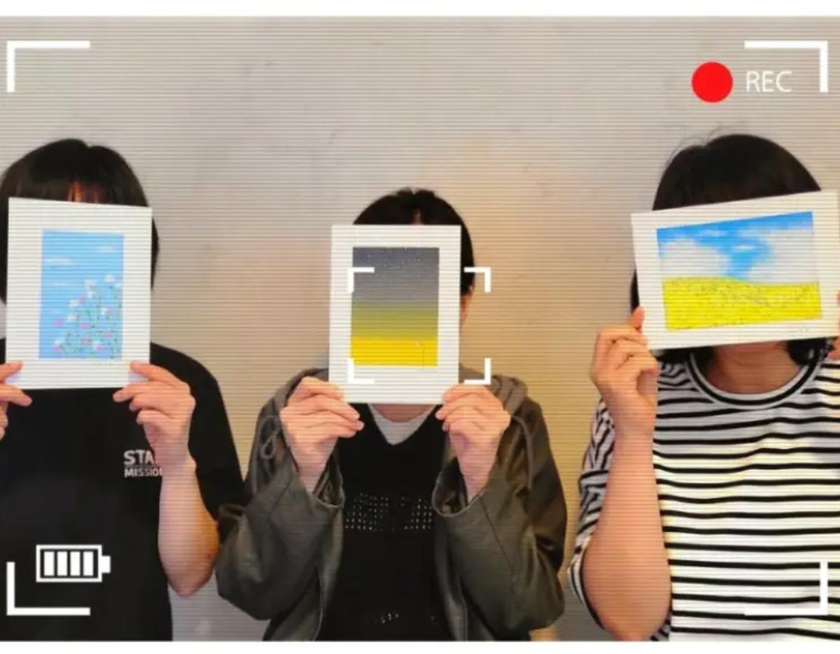 three participants seated side-by-side holding up finished A4 oil pastel landscapes covering their faces, camera overlay icons visible